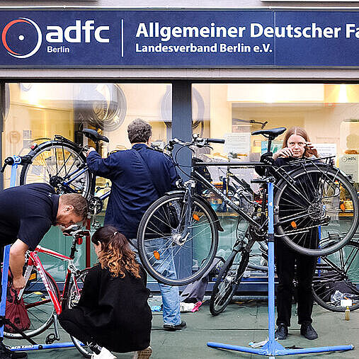 Immer bestens informiert Mehrere Menschen reparieren Fahrräder auf Ständern vor einem Gebäude mit ADFC‑Berlin‑Schild.