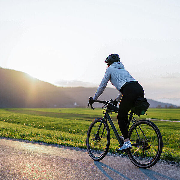 Entspannt und sicher unterwegs Eine Person fährt mit dem Fahrrad auf einer ruhigen Straße durch eine weite Wiesenlandschaft, die Sonne steht tief am Himmel.