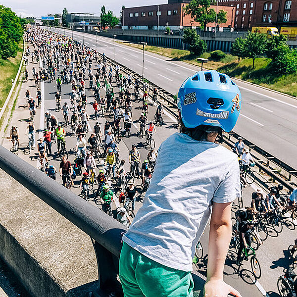 Bewegung in die Zukunft Ein Kind mit Helm steht auf einer Brücke und blickt auf eine volle Straße, auf der viele Menschen mit Fahrrädern fahren.