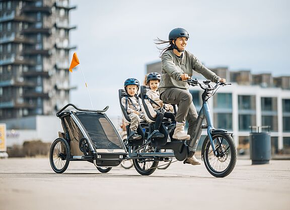 Familienlogistik: Longtail mit zwei Kindersitzen plus Anhänger Frau fährt ein Lastenrad Longtail, auf dem langen Gepäckträger hinten sitzen zwei Kinder in je einem Kindersitz. Das Fahrrad zieht zudem einen Kinderfahrradanhänger.