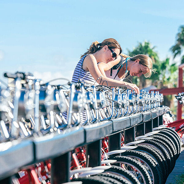Mobil auch ohne eigenes Rad Zwei Personen beugen sich über eine lange Reihe ausgeliehener Fahrräder und prüfen die Räder an einer Station.