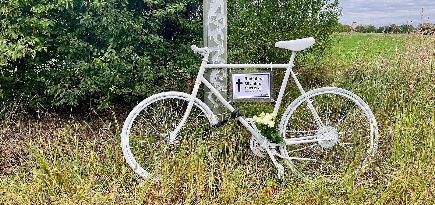 weißes Geisterrad mit Schild und weißen Rosen am Wegrand