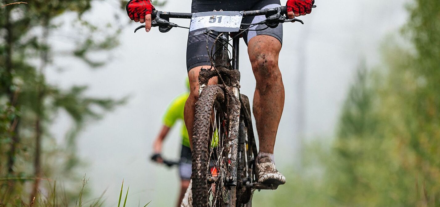 Ein  Mountainbike-Fahrer, verschmutzt und mit Startnummer am Lenker, sichtbar bis knapp oberhalb des Lenkers, fährt bergauf. Im Hinergrund ist ein weiterer Fahrer zu sehen. 