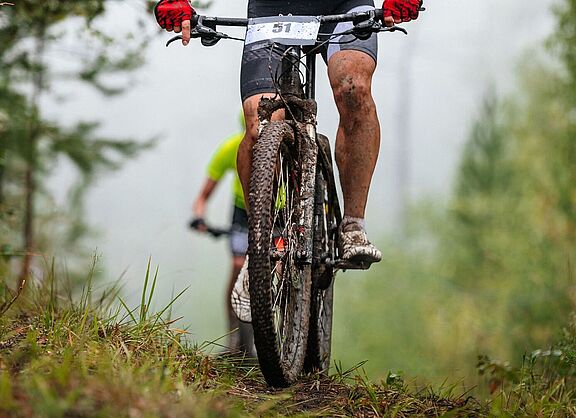 Ein  Mountainbike-Fahrer, verschmutzt und mit Startnummer am Lenker, sichtbar bis knapp oberhalb des Lenkers, fährt bergauf. Im Hinergrund ist ein weiterer Fahrer zu sehen. 