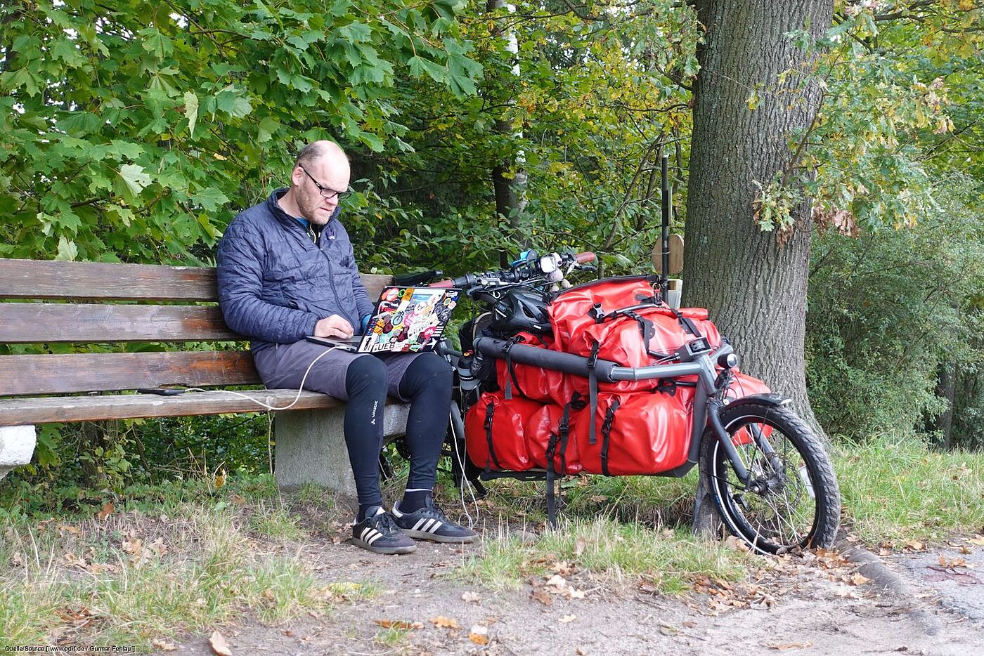 Gunnar Fehlau sitzt auf einer Bank und arbeitet am Laptop. Sein Lastenrad steht neben ihm. 