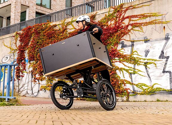 Chike Kurve Person mit Helm fährt ein dreirädriges Lastenrad mit großer Transportbox vor einer bewachsenen Wand.