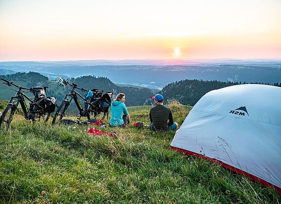 Radreise und Zelten Zwei Radreisende sitzen in der Dämmerung neben einem Zelt im Grünen und blicken auf einen Höhenzug in der Ferne.