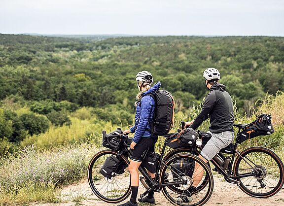 Bikepacking Artikel Zwei Radfahrende mit Gravelbikes und Gepäck stehen auf einer Anhöhe und blicken auf einen weiten Wald.