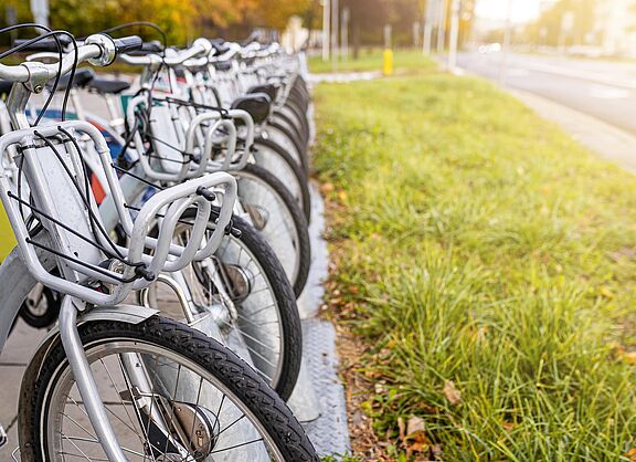 Öffentliche Leihfahrräder an einer Bike-Sharing-Station Eine Reihe von öffentlichen Leihfahrrädern mit silbernen Rahmen und grauen Körben am Lenker steht ordentlich nebeneinander an einer stationsgebundenen Verleihstation. Die Fahrräder sind auf einem gepflasterten Gehweg abgestellt, neben dem eine Grünfläche verläuft.