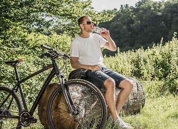 Radfahrer macht Pause im Grünen Radfahrer mit Sonnenbrille trinkt Wasser aus einer Flasche während einer Pause auf einem Baumstamm. Sein schwarzes Mountainbike steht daneben.