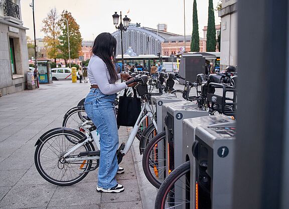 Frau entleiht Fahrrad an Bike-Sharing-Station Eine junge Frau mit langen dunklen Haaren, weißem Oberteil, Jeans und schwarzer Umhängetasche steht an einer Bike-Sharing-Station und entnimmt ein weißes Leihfahrrad. Die Station verfügt über mehrere nummerierte Stellplätze mit grauen Dockingelementen. Im Hintergrund sind ein belebter Stadtplatz, historische Gebäude, Passanten und ein Bahnhofsgebäude zu sehen.