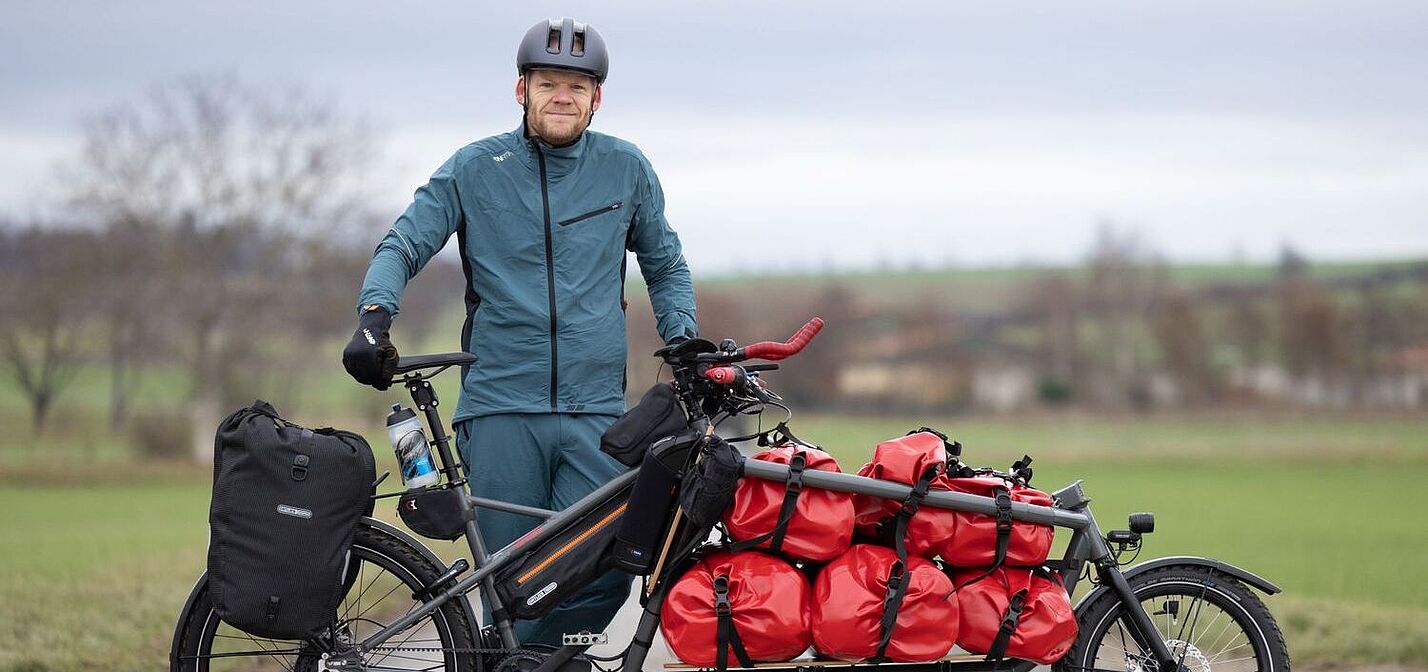 Gunnar Fehlau steht in Fahrradkleidung, vor ihm sein Lastenrad, mit dem er auf Workpacking-Tour ist. 