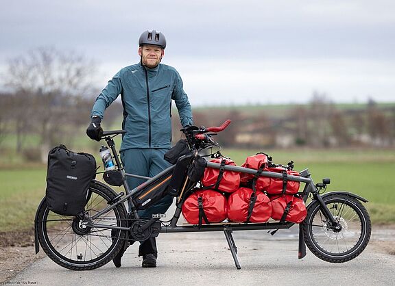 Gunnar Fehlau steht in Fahrradkleidung, vor ihm sein Lastenrad, mit dem er auf Workpacking-Tour ist. 