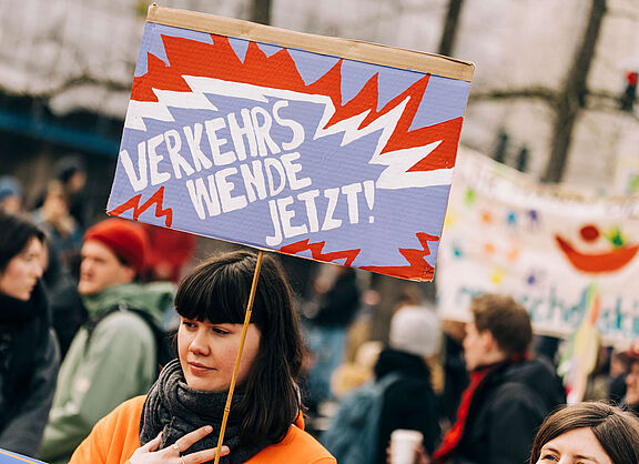 Junge Frau auf einer Demo hält ein Schild in die Luft, auf dem steht "Verkehrswende Jetzt!" 