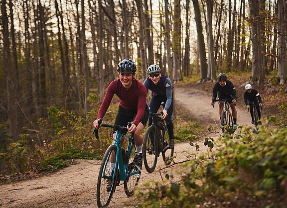 Eine Gruppe von Frauen und Männern fahren mit Gravelbikes hintereinander auf einem Waldweg. 