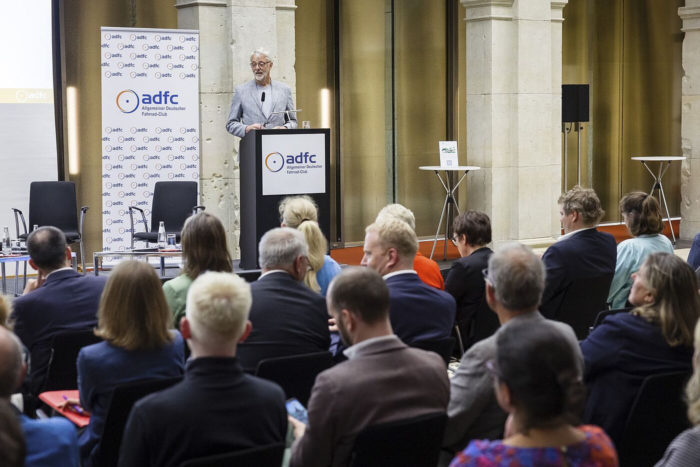 ADFC-Bundesvorsitzender Frank Masurat am Pult spricht zu den Zuschauenden im Vordergrund. 