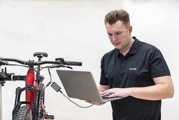 Mann mit Laptop steht neben einem Fahrrad. Das Laptop ist an das Fahrrad angeschlossen. Der Mann liest die elektronisch verfügbaren Daten mit seinem Laptop vom Fahrradmotor aus.