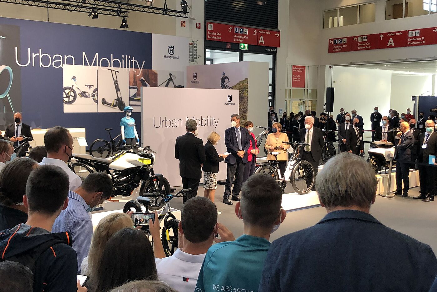 Angela Merkel mit Begleitern auf einem Messestand mit Fahrrädern von Zuschauern umringt. 