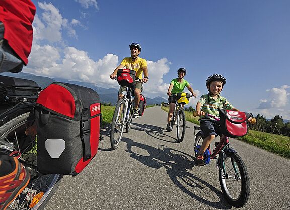 Sommerliche Familienradtour auf dem Land Eine vierköpfige Familie fährt mit Helmen und Packtaschen auf Fahrrädern eine schmale Landstraße entlang; vorn ein Kind auf einem kleinen Rad, dahinter zwei Erwachsene und ein weiteres Fahrrad im Anschnitt links, umgeben von Wiesen, Bergen und blauem Himmel.
