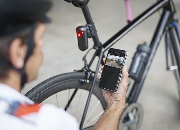 Rücklicht Garmin Varia RCT715 Mann kniet mit Smartphone in der Hand vor einen Rennrad mit rot leuchtendem Rücklicht an Sattelstütze. Das Rücklicht Garmin Varia RCT715 mit integrierter Kamera zeichnet permanent auf und speichert Daten aber nur bei einem erkannten Unfall.