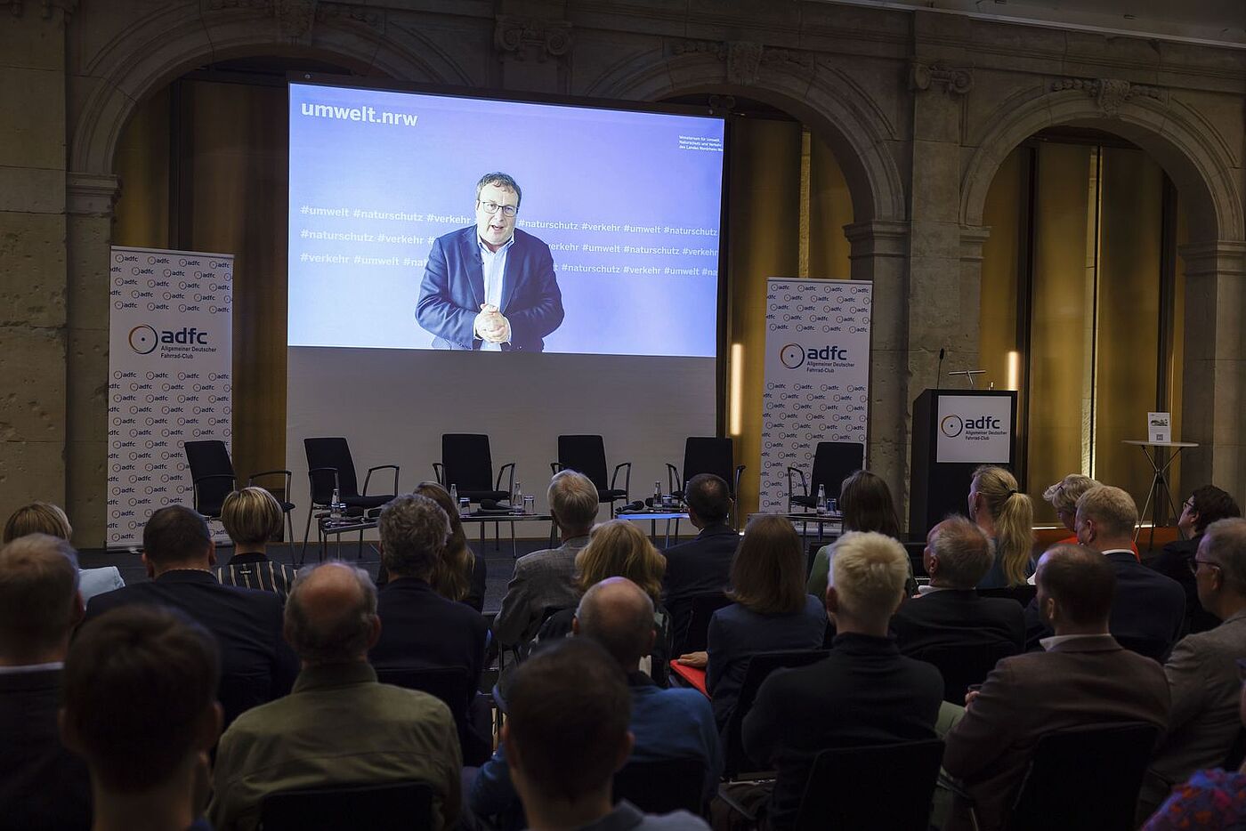 Auf der Videoleinwand ist Oliver Krischer, Minister für Umwelt, Naturschutz und Verkehr des Landes Nordrhein-Westfalen, zu sehen, der ein Grußwort spricht. 