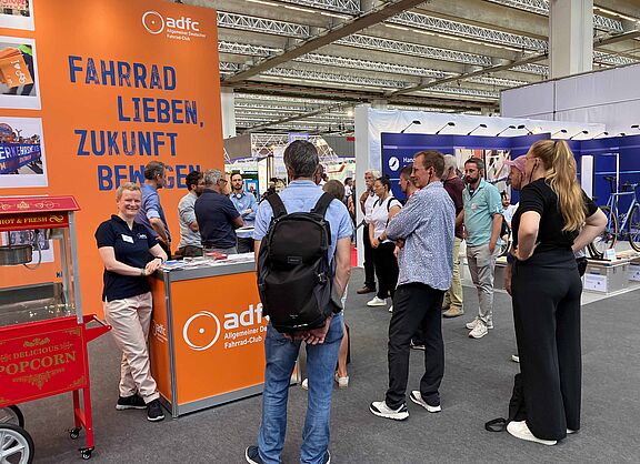 Eurobike 2025 Ein Messestand des ADFC in einer großen Halle. Vor einer orangen Rückwand mit dem blauen Slogan „Fahrrad lieben, Zukunft bewegen“ steht eine lächelnde Frau im dunkelblauen ADFC-Shirt an einer Theke. Mehrere Menschen stehen davor und dahinter in Gruppen zusammen und unterhalten sich. Im Vordergrund links ist eine rote Popcornmaschine zu sehen.