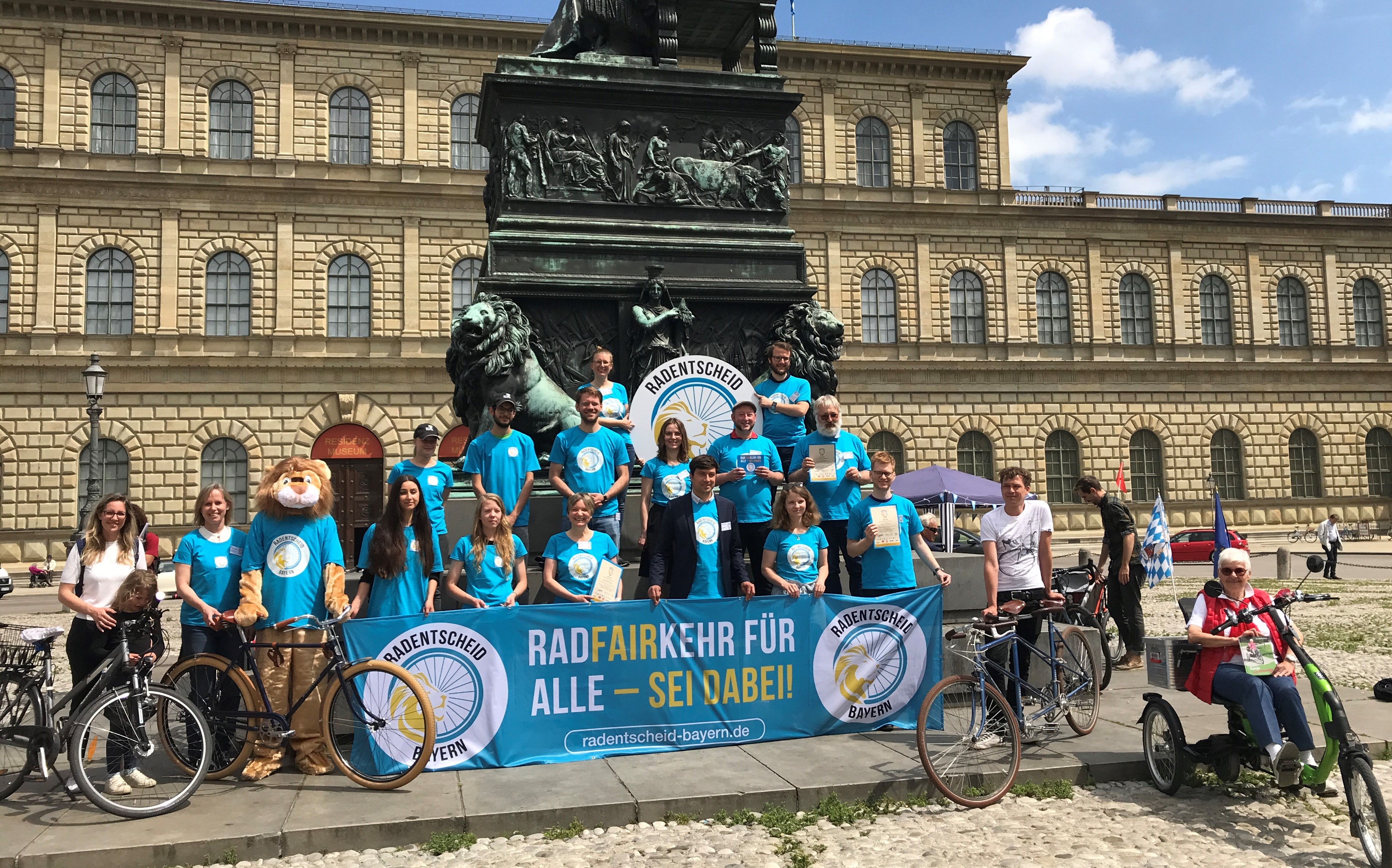 Etwa 15 Personen in blauen Aktions-T-Shirts präsentieren auf einem Platz in München das Transparent des Radentscheids Bayern. 