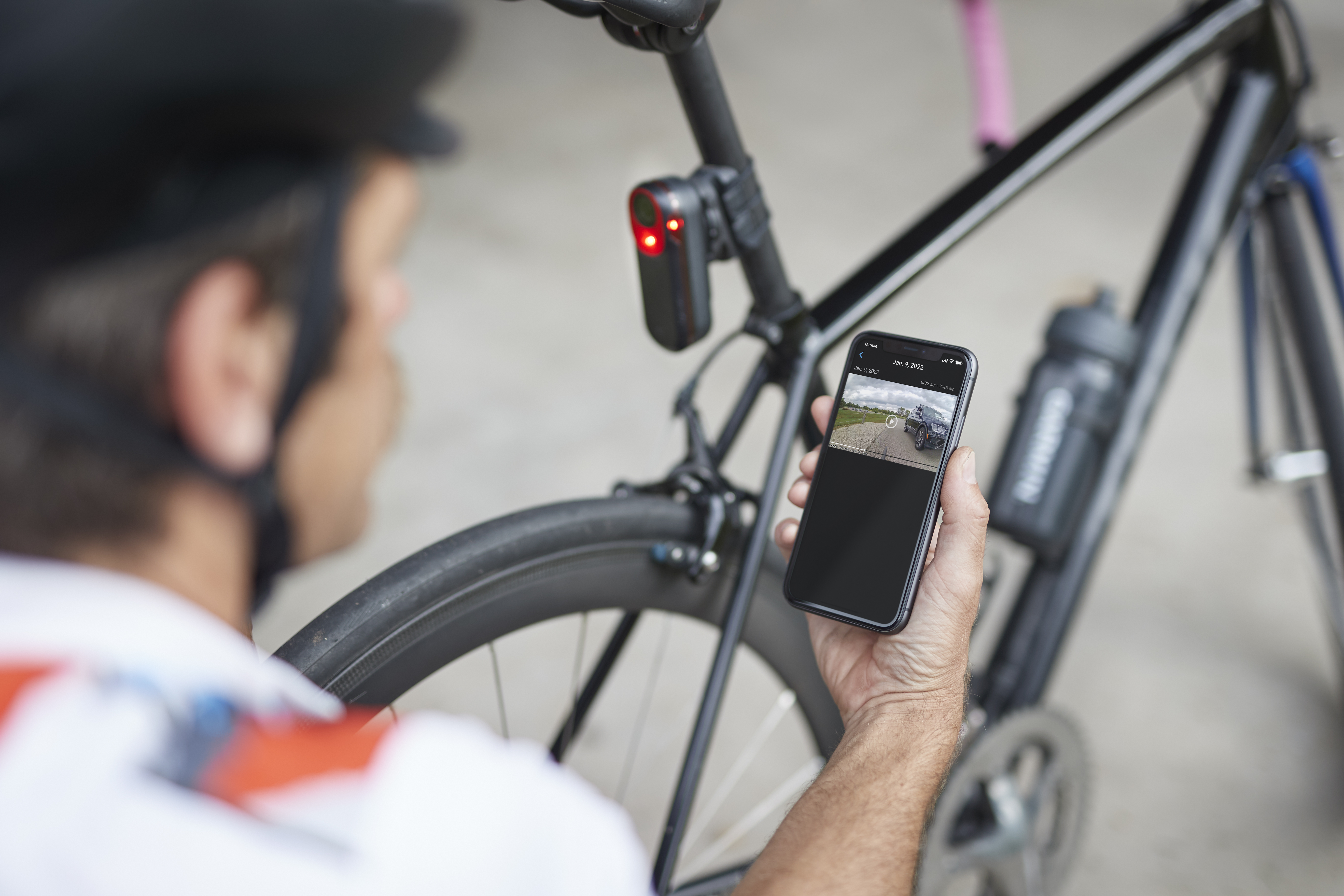 Mann kniet mit Smartphone in der Hand vor einen Rennrad mit rot leuchtendem Rücklicht an Sattelstütze. Das Rücklicht Garmin Varia RCT715 mit integrierter Kamera zeichnet permanent auf und speichert Daten aber nur bei einem erkannten Unfall.  